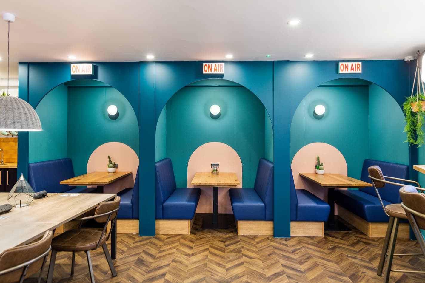 Modern teal alcove booths with blue benches, wood tables, and overhead “ON AIR” signs.